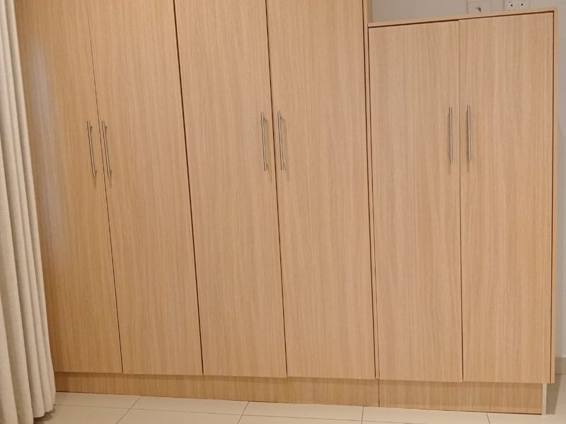A clean bedroom featuring light wood-finish built-in cupboards and neutral tiled flooring, offering ample storage space.