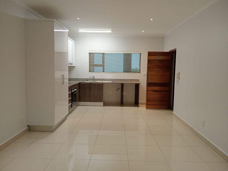 An open-plan kitchen and living area featuring modern cabinetry, granite countertops, built-in oven, and neutral ceramic floor tiles in a clean, vacant space.
