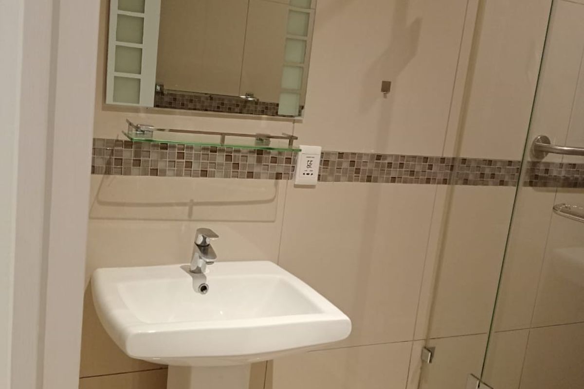 A clean, modern bathroom featuring a pedestal basin, glass shower enclosure, and neutral ceramic tiling with a decorative mosaic border.