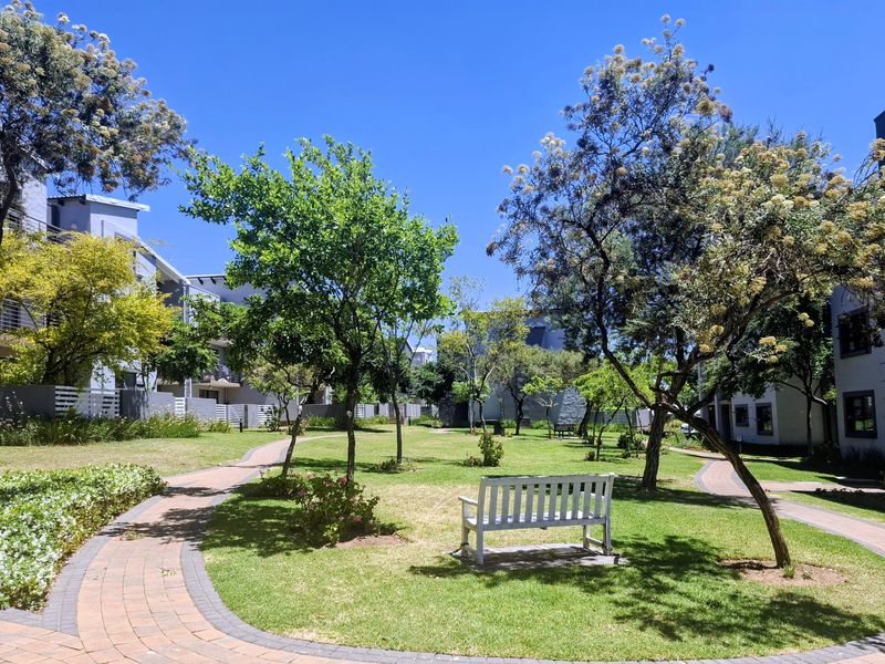 A well-maintained communal garden area featuring paved walkways, lush green lawns, and mature trees within an apartment complex setting.