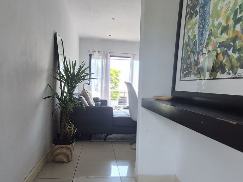 A bright hallway featuring neutral tiled flooring and recessed downlighting, leading towards an open-plan living area with natural light.