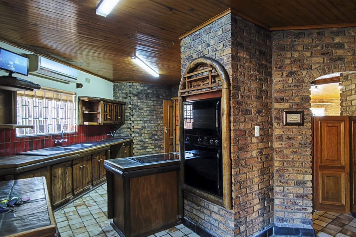 A rustic kitchen featuring exposed brick walls, timber ceiling, built-in oven, and ample wooden cabinetry with tiled flooring.