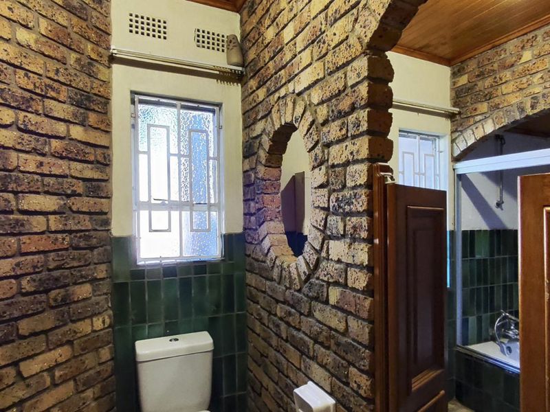 A rustic bathroom featuring exposed brick walls, timber ceiling, and a combined bath and toilet area with dated green tiling.