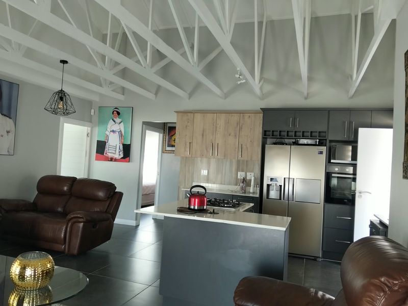 Modern open-plan living area featuring a contemporary kitchen with two-tone cabinetry, stone countertops, built-in appliances, and striking exposed white roof trusses.