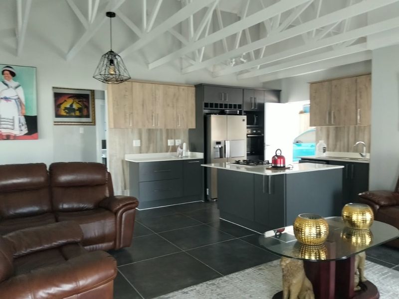 Modern open-plan living area featuring a contemporary kitchen with a central island, stone countertops, built-in appliances, and striking exposed white roof trusses.