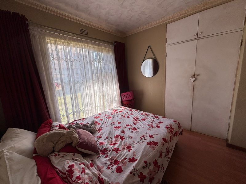 A bedroom featuring built-in cupboards, laminate flooring, and a large window with security gate, offering a functional space with potential for cosmetic updates.