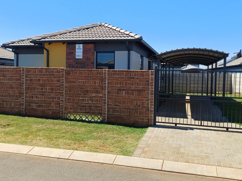 Front view of a modern house featuring a brick boundary wall, paved driveway, and a covered carport with a metal gate.