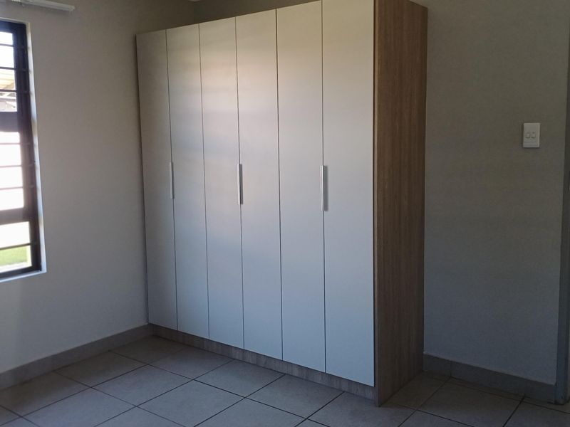 A clean, empty bedroom featuring neutral tiled flooring, a modern built-in wardrobe with light-toned doors, and a window fitted with security bars.