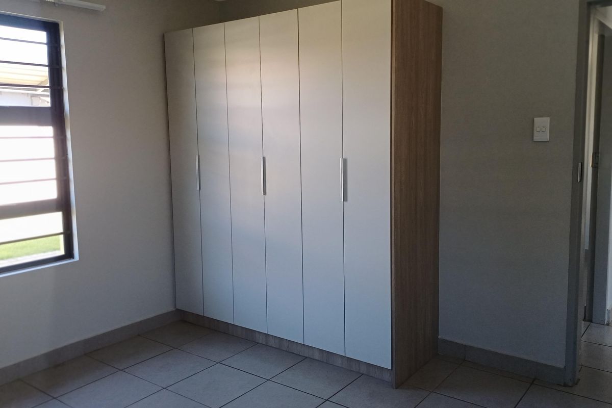 A clean, empty bedroom featuring neutral tiled flooring, a modern built-in wardrobe with light-toned doors, and a window fitted with security bars.