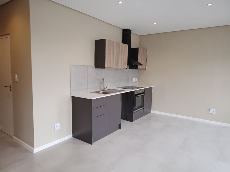 Modern, compact kitchen featuring sleek dark cabinetry, stone countertops, integrated oven, hob, and extractor fan, set against a neutral tiled floor.
