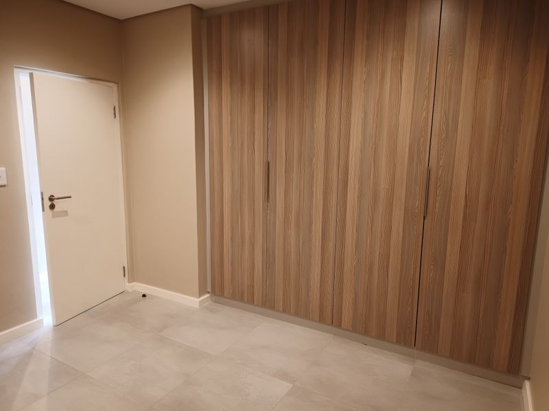 A clean, modern bedroom featuring neutral wall tones, large-format floor tiles, and extensive floor-to-ceiling built-in wooden cabinetry.