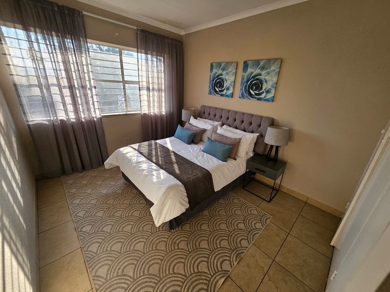 A well-lit bedroom featuring tiled flooring, neutral wall tones, and a large window fitted with security bars and curtains.