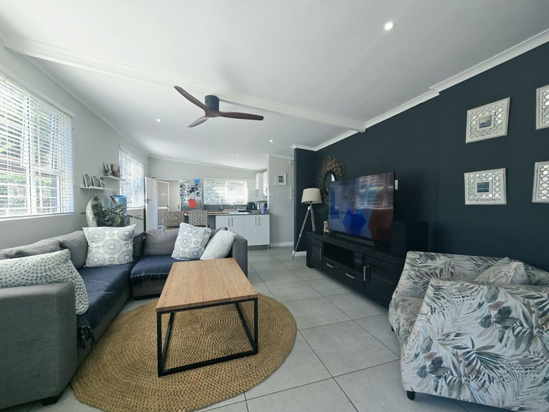 An open-plan living area featuring tiled flooring, a ceiling fan, and recessed lighting, seamlessly connecting to a functional kitchen space with built-in cabinetry.