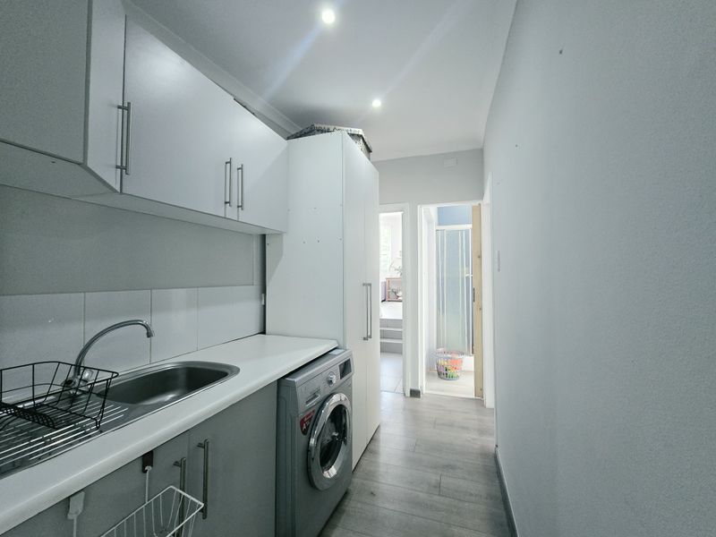 A functional laundry area featuring built-in cabinetry, a stainless steel sink, and space for a washing machine, with modern laminate flooring.
