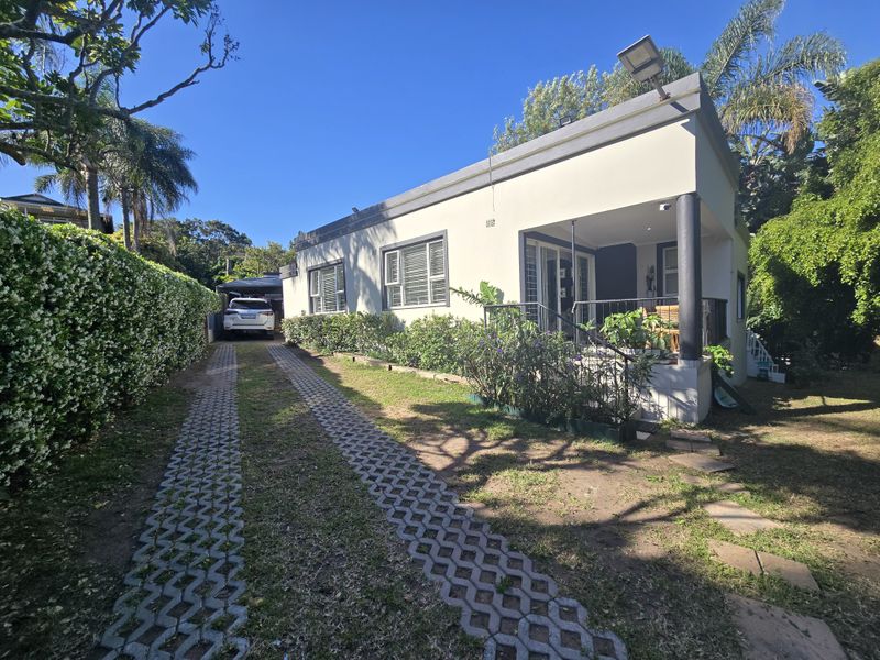 Front exterior view of the house featuring a paved driveway, covered patio area, and established garden hedging.