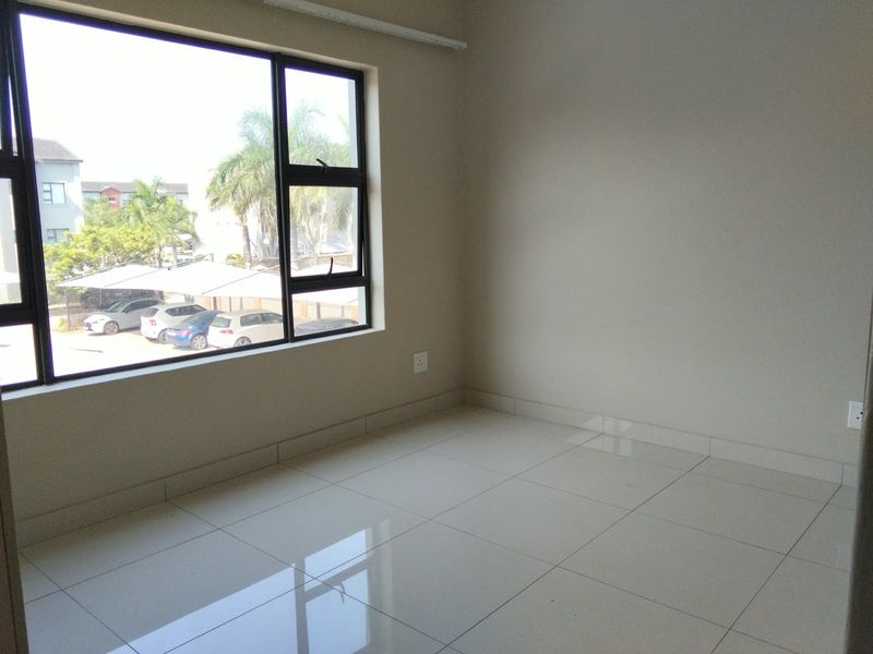 A bright, empty bedroom featuring neutral tiled flooring, clean walls, and a large window providing natural light and a view of the parking area.