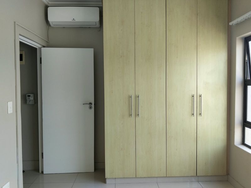 A clean, empty bedroom featuring light wood-finish built-in cupboards, tiled flooring, and a wall-mounted air conditioning unit.