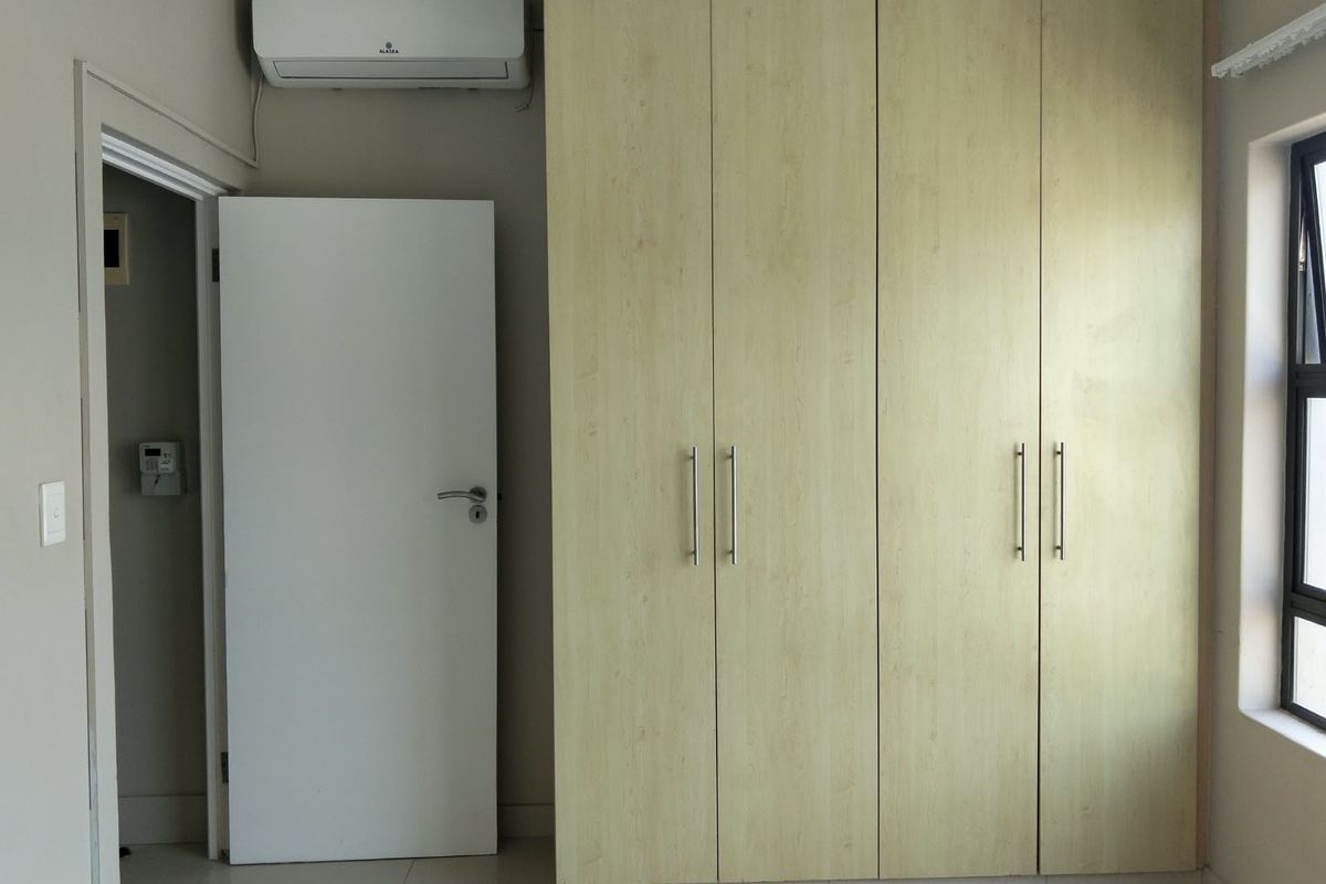 A clean, empty bedroom featuring light wood-finish built-in cupboards, tiled flooring, and a wall-mounted air conditioning unit.