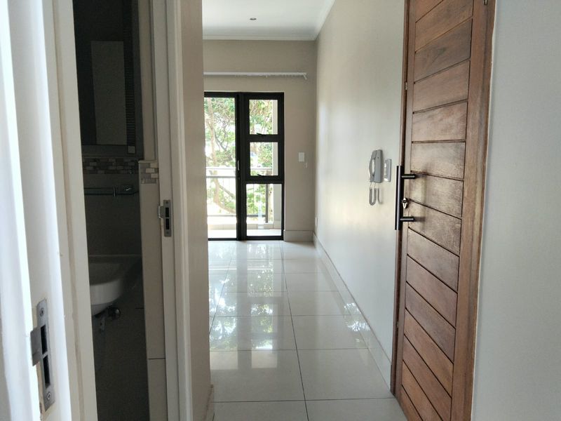 A bright, tiled hallway featuring a wooden door, intercom system, and a sliding glass door leading to an outdoor balcony area.