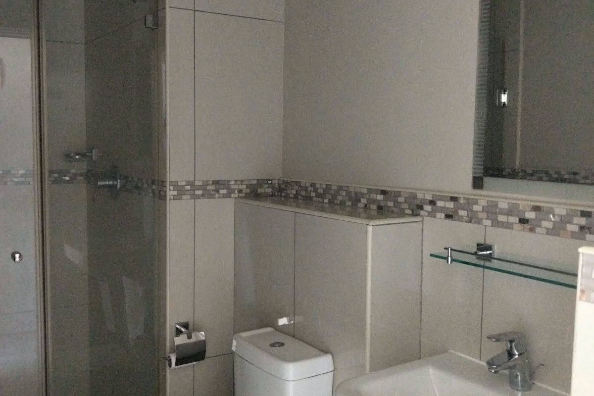 A clean, modern bathroom featuring a pedestal basin, toilet, and a glass-enclosed shower with neutral wall tiling and a decorative mosaic border.