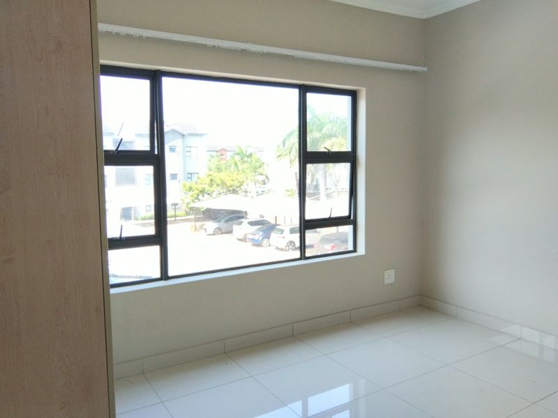 A bright, empty bedroom featuring neutral tiled flooring, built-in cupboards, and large aluminium-framed windows overlooking the complex parking area.