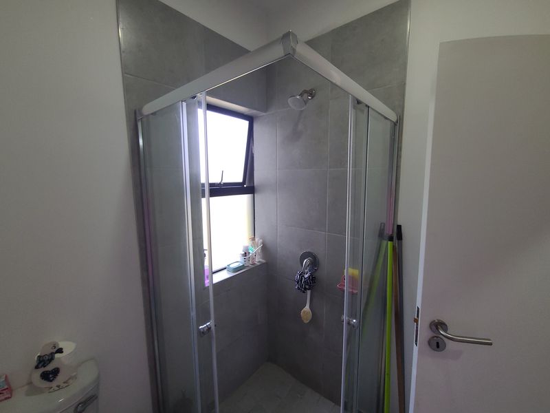 A functional bathroom featuring a corner shower enclosure with grey tiling, a window for natural light, and a standard toilet.