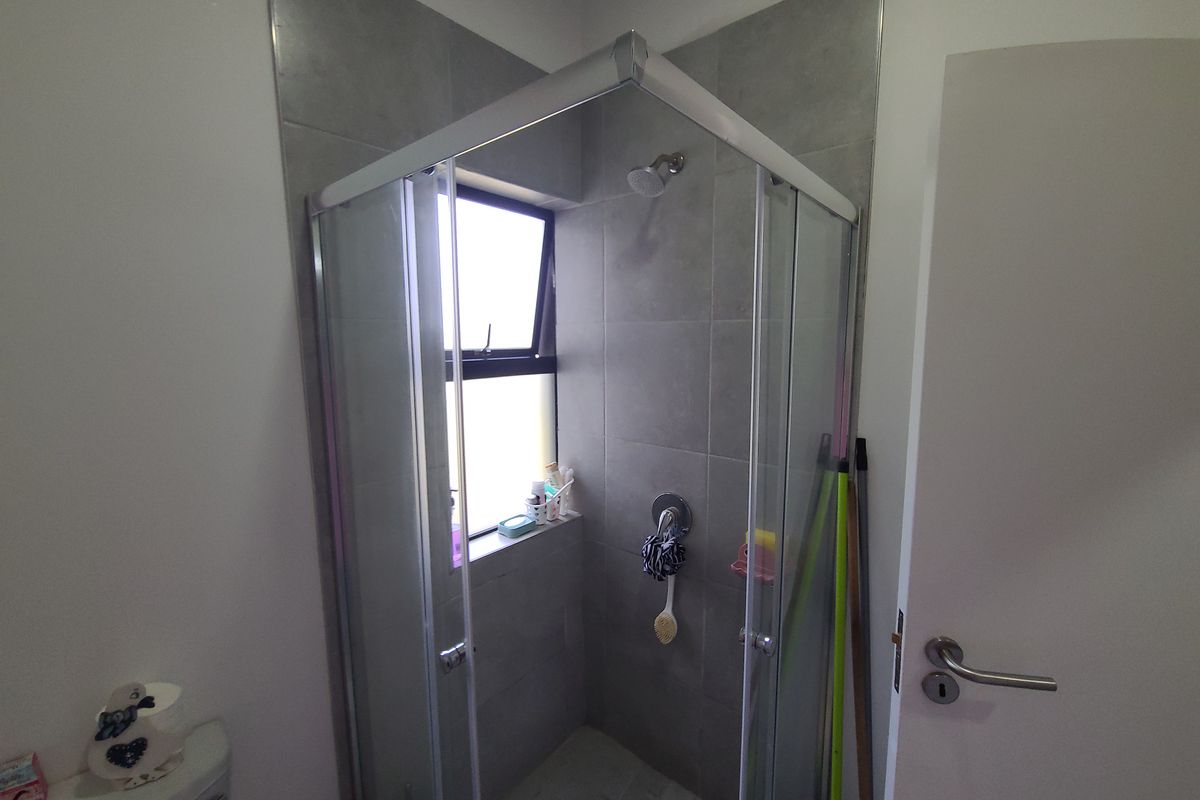 A functional bathroom featuring a corner shower enclosure with grey tiling, a window for natural light, and a standard toilet.