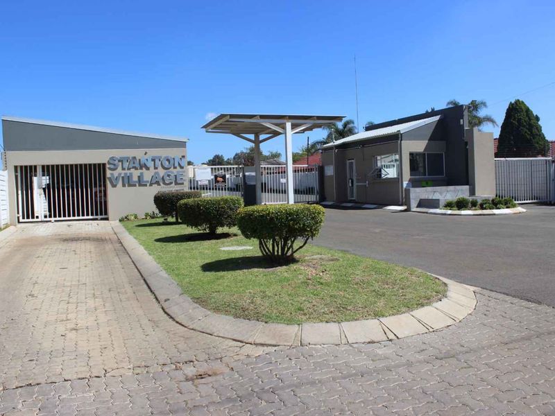 The entrance to Stanton Village featuring a secure gated access point, guard house, paved driveway, and well-maintained landscaped garden areas.