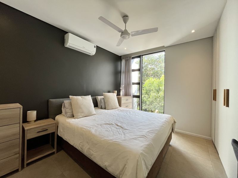 A modern bedroom featuring tiled flooring, built-in cupboards, a ceiling fan, and a split-system air conditioner, with natural light from a large window.