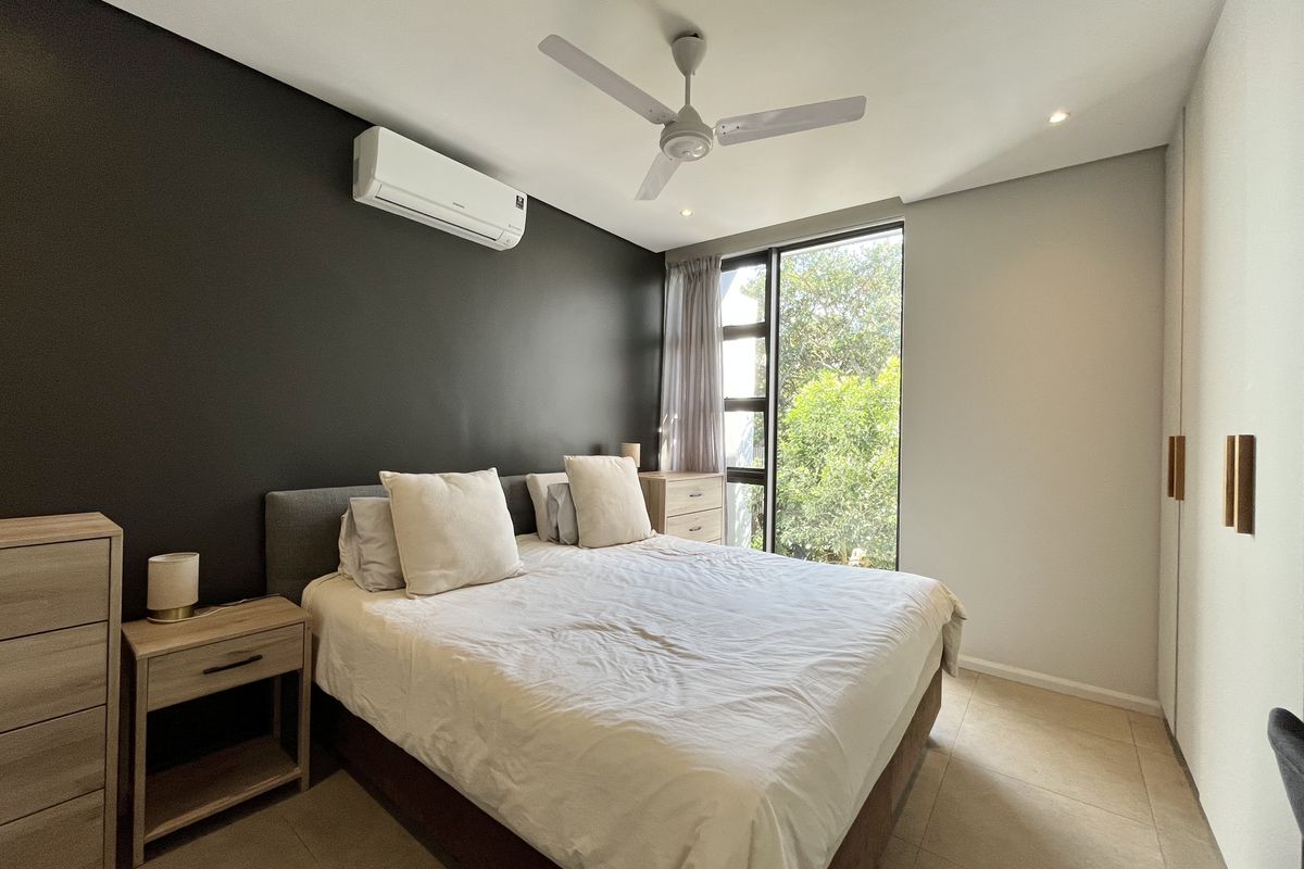 A modern bedroom featuring tiled flooring, built-in cupboards, a ceiling fan, and a split-system air conditioner, with natural light from a large window.