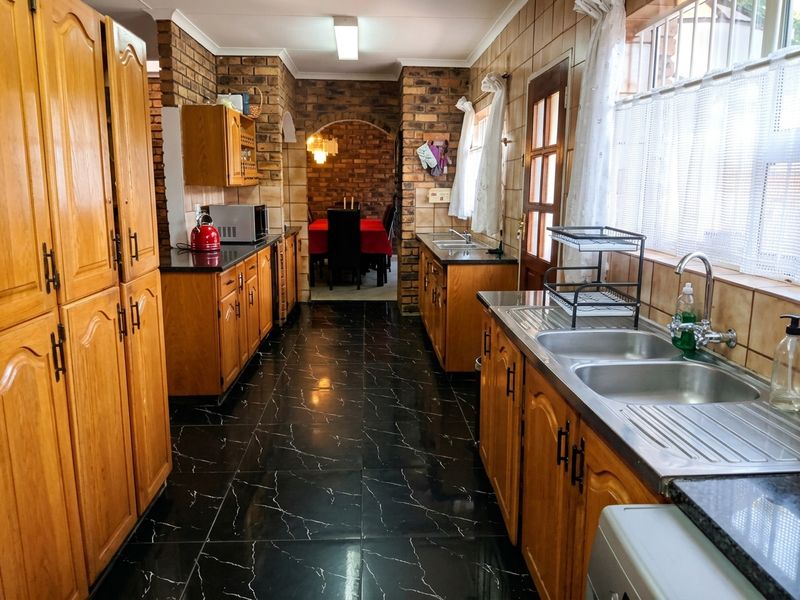 A functional kitchen featuring wooden cabinetry, double stainless steel sink, and striking black tiled flooring, with an adjacent dining area visible through an exposed brick archway.