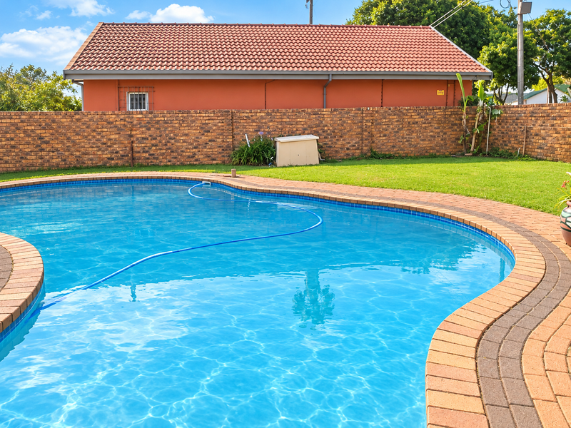 A well-maintained swimming pool area featuring a curved paved surround, a lush lawn, and a solid brick boundary wall.