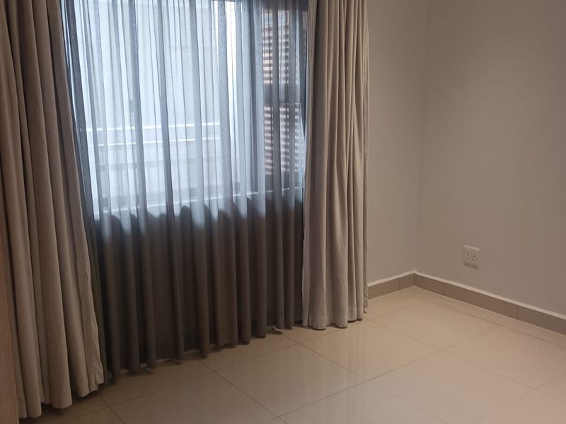 A bright, empty bedroom featuring neutral tiled flooring, recessed ceiling lighting, and large windows dressed with floor-to-ceiling curtains.