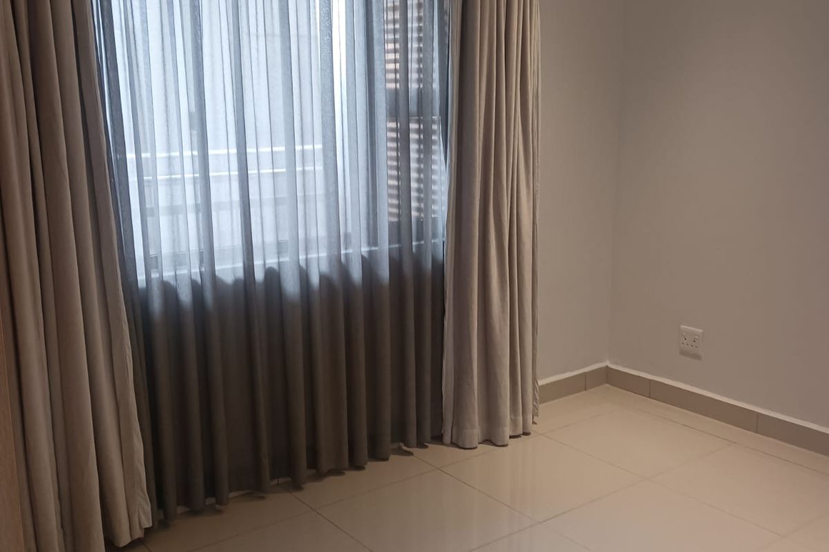 A bright, empty bedroom featuring neutral tiled flooring, recessed ceiling lighting, and large windows dressed with floor-to-ceiling curtains.