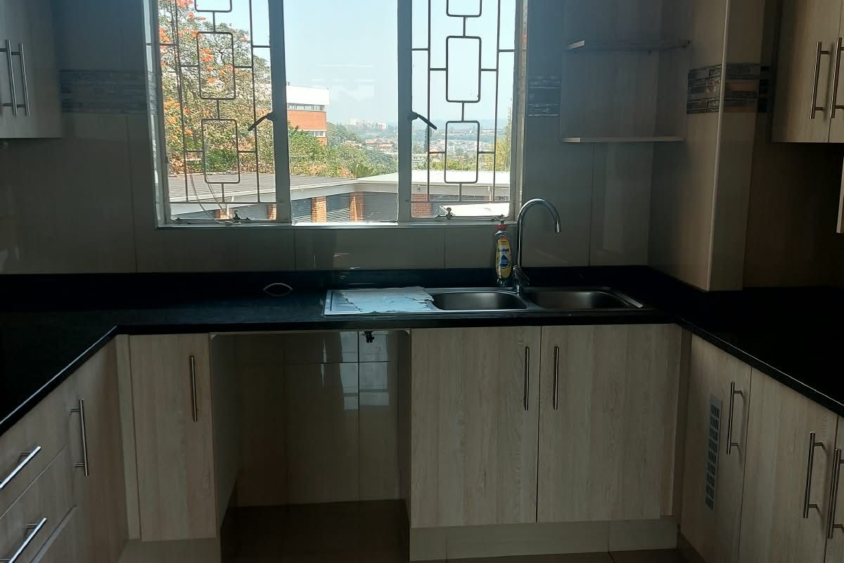 A functional kitchen featuring light wood-finish cabinetry, dark granite countertops, a double basin sink, and large windows with security bars.