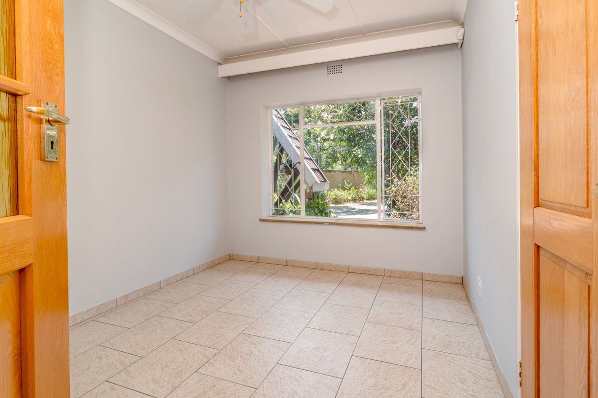 A bright, empty bedroom featuring neutral tiled flooring, a ceiling fan, and a large window with security bars overlooking the garden.