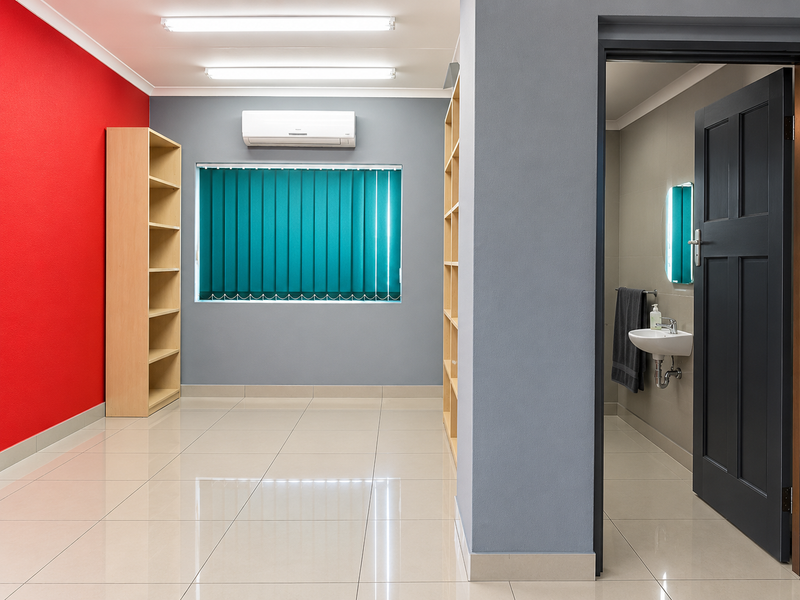 A bright, modern office space featuring tiled floors, built-in shelving, air conditioning, and an adjoining private washroom with a wall-mounted basin.