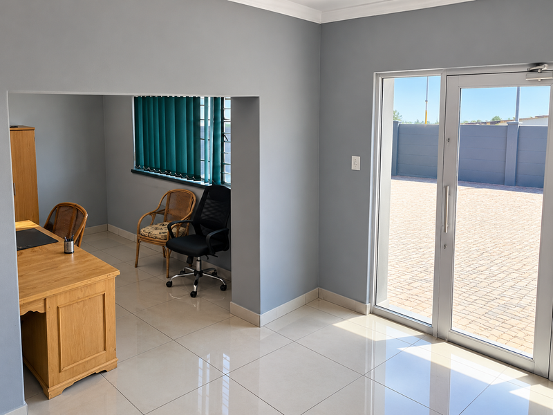A bright office space featuring neutral grey walls, tiled flooring, and a large aluminium sliding door providing direct access to the paved outdoor area.