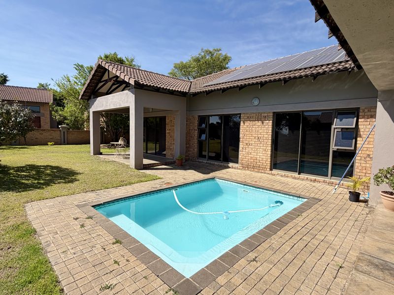 Rear exterior view featuring a swimming pool, paved patio area, and covered veranda, set against a brick house with solar panels.