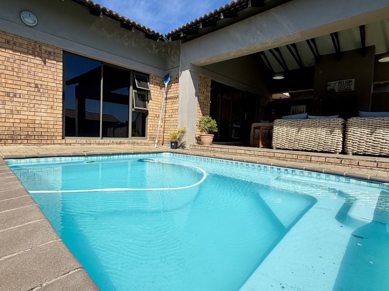 A well-maintained swimming pool area featuring a covered patio with exposed beams, brick paving, and direct access to the home via sliding glass doors.