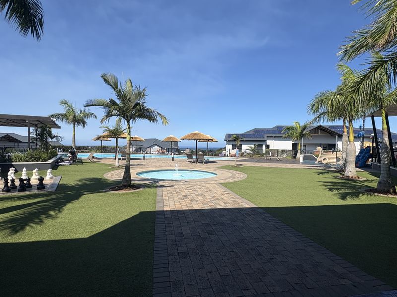 A well-maintained communal pool area featuring a central water fountain, paved pathways, artificial turf, and established palm trees within a residential complex.