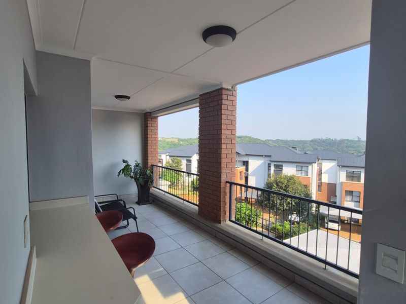 A spacious covered balcony featuring tiled flooring, brick pillars, a metal balustrade, and a built-in counter, offering pleasant views of the surrounding residential complex.