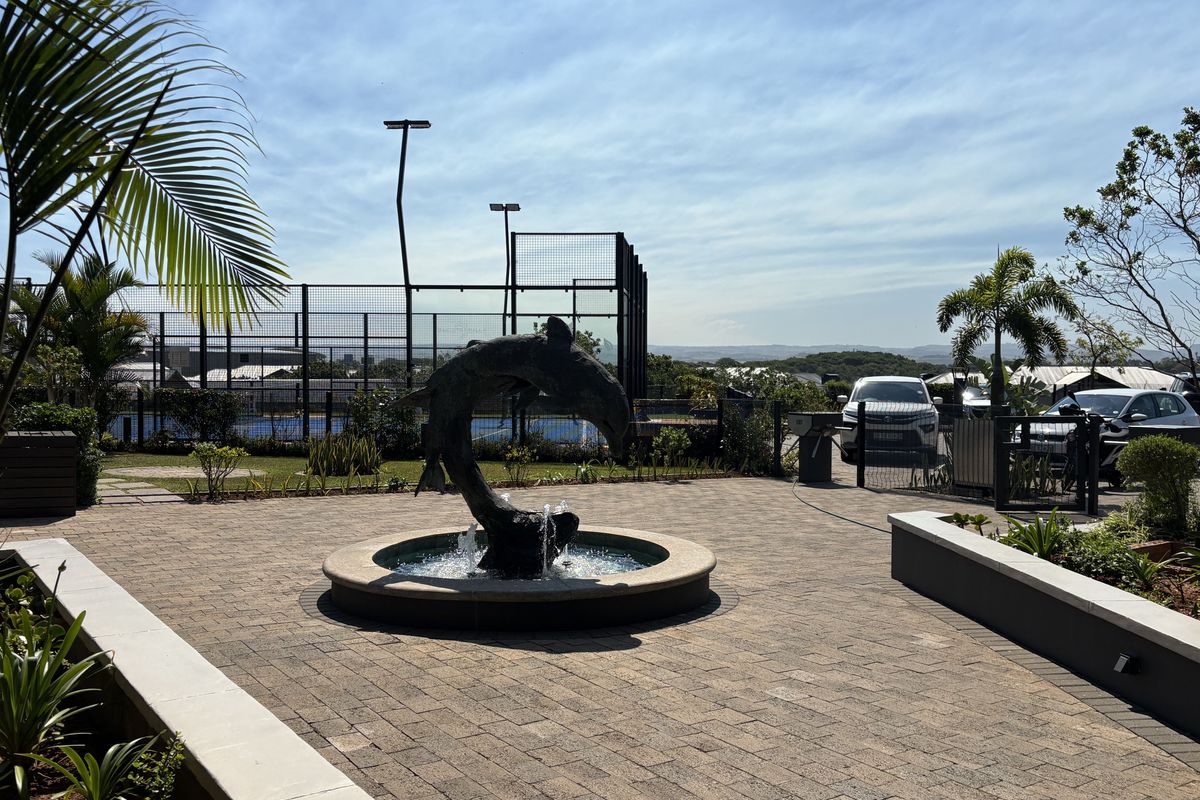 A well-maintained paved courtyard featuring a central water fountain, landscaped garden beds, and adjacent sports court facilities.