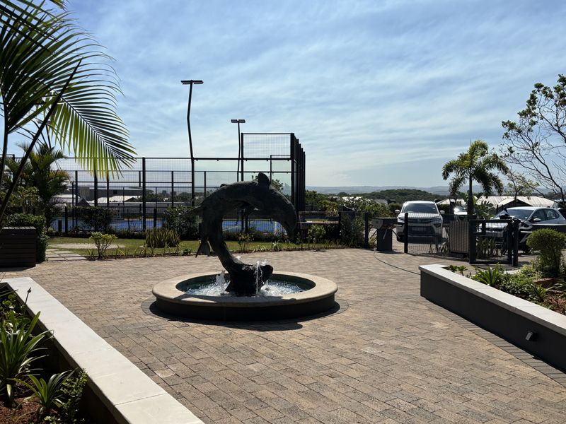 A well-maintained paved courtyard featuring a central water fountain, landscaped garden beds, and adjacent sports court facilities.