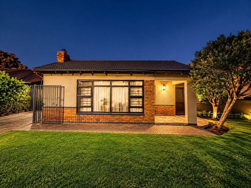 A well-maintained rear exterior view of a brick house featuring a paved patio, manicured lawn, and secure metal gate at twilight.
