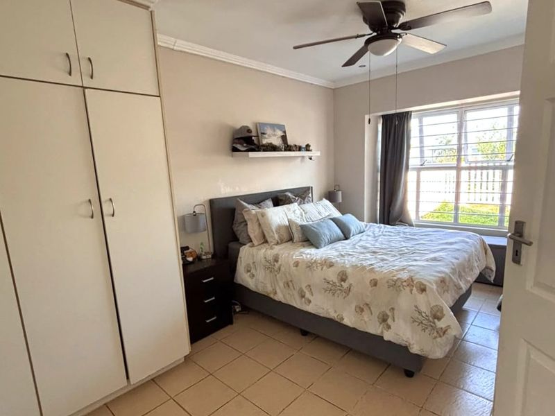 A functional bedroom featuring built-in cupboards, tiled flooring, and a ceiling fan, with natural light provided by a window fitted with security bars.