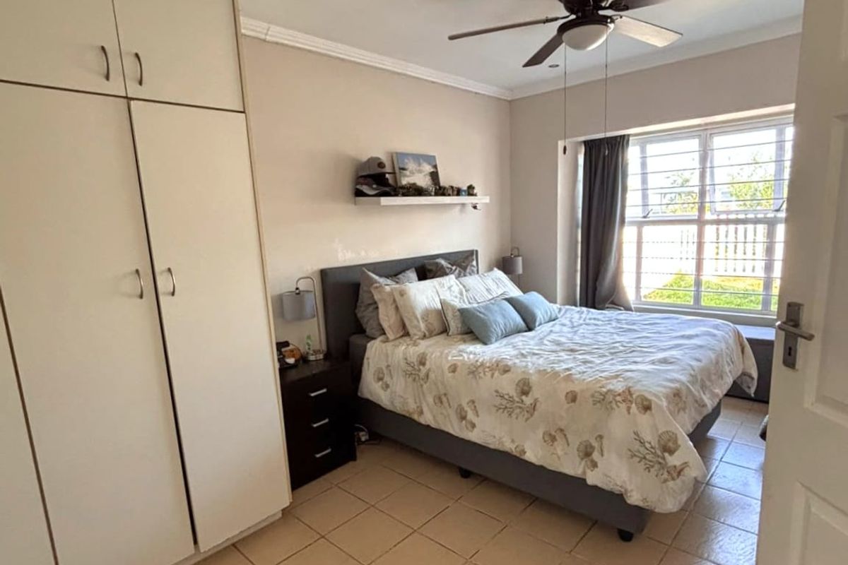 A functional bedroom featuring built-in cupboards, tiled flooring, and a ceiling fan, with natural light provided by a window fitted with security bars.