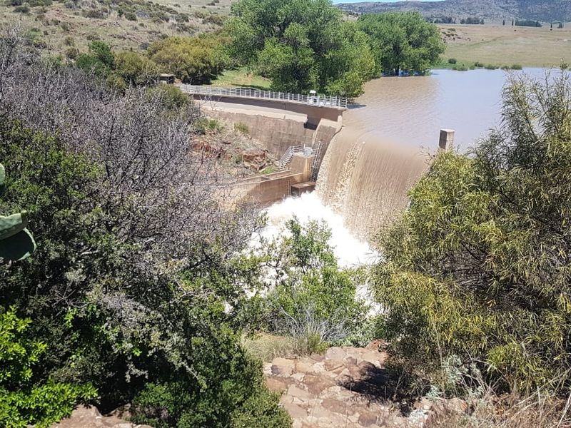 A scenic view of a concrete dam wall with water flowing over the spillway, set within a natural rural landscape.