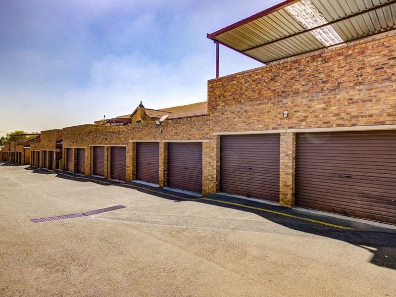 A row of secure garages featuring face brick construction and roller shutter doors, situated along a paved driveway within the townhouse complex.