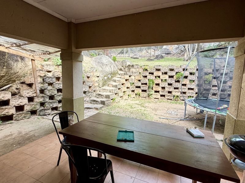 Covered patio area featuring tiled flooring and concrete pillars, overlooking a terraced garden with stone retaining walls and natural rock features.